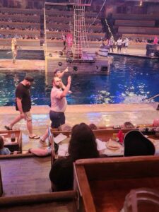 People walking by a water stage with a pirate ship set, surrounded by seated audience members enjoying food and drinks during a live performance.