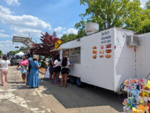 People waiting at a white food truck called Harley's Concession during an outdoor event, with a rack of stuffed toys and a performance stage nearby.