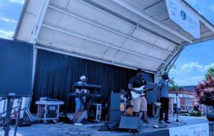 A band performs on an outdoor stage under a white canopy. Musicians play guitar, keyboard, and drums, with a vocalist singing into a microphone. Blue sky and trees in background.