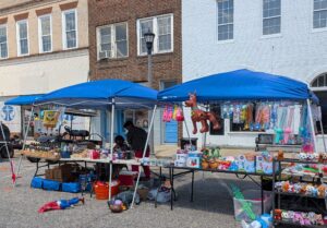 Outdoor street market stall under blue tents, selling toys, inflatables, plush animals, and novelty items. People are behind the tables, and colorful buildings are in the background.