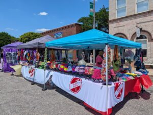 Outdoor market with colorful crocheted hats, bags, and crafts displayed under tents on a sunny day, with people browsing in the background near brick buildings.