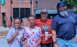 Four people pose outdoors in front of a bank building, two children eating ice cream and two adults smiling, on a sunny day.