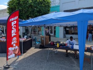 A street vendor sits under a blue canopy next to a red "Fried Pork Skin" flag, with cooking equipment and condiments on the table.