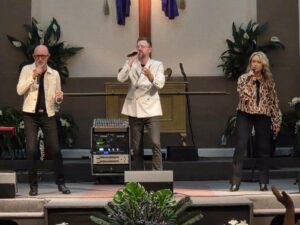 Three people sing on a church stage, each holding a microphone, with a cross and plants in the background.