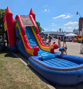 A colorful inflatable slide at an outdoor event, with children playing and adults standing nearby under a clear blue sky.