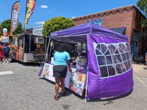 Outdoor food festival with a seafood food truck, people waiting, and a purple tent selling baked goods on a sunny day. Brick building and blue sky in the background.