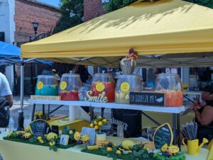 A lemonade stand at an outdoor festival with colorful drinks in large jars, yellow decorations, and signs with cheerful messages under a bright yellow canopy.