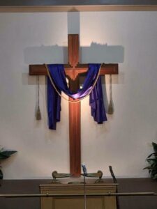A wooden cross draped with a purple cloth and gold rope is mounted on a church wall, with a microphone stand below.