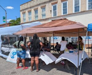 People gather under tents at a community street festival, with tables covered in white cloth and a sign reading "Community Welcome" visible in the foreground.