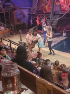 Performers in pirate costumes interact with audience members at an indoor pirate-themed dinner show with a large ship and water stage.