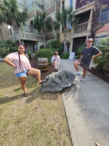 Three people posing with one foot on a large rock, outdoors near palm trees and a building with a rustic facade.