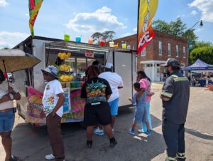 People waiting in line at a lemonade and drink stand during a sunny outdoor event, with colorful drinks displayed and a brick building in the background.