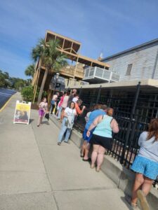 People stand in a line outside a building on a sunny day, with a sign advertising an event visible on the sidewalk.