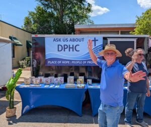 Two men stand in front of a booth with a "ASK US ABOUT DPHC" sign, informational materials, and a potted plant, outdoors on a sunny day.