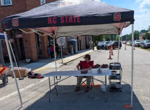 Person sitting under NC State tent at outdoor market, selling strawberries in baskets, with cars parked on the street and people gathered nearby on the sidewalk.