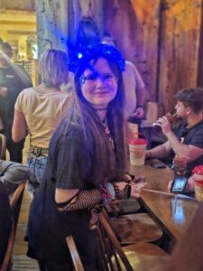 A young woman with long hair wears blue glowing headgear, face gems, and fishnet gloves, smiling at a lively restaurant surrounded by people.