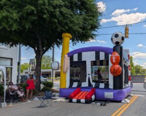 Inflatable bounce house decorated with large sports balls on a city street, with two women sitting and standing nearby under a tree on a sunny day.