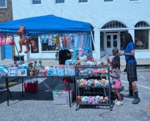 A man and two children browse a street vendor's toy stall, featuring stuffed animals, inflatable toys, and bubble guns under a blue canopy on a sunny day.