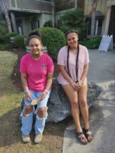 Two young women sit on a large rock outside, smiling. One wears a pink shirt and ripped jeans, the other a pink top and shorts.