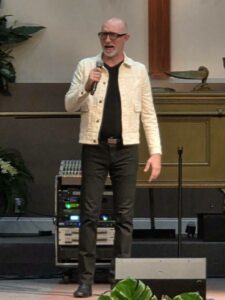 A man with glasses and a white jacket is speaking into a microphone on stage, standing in front of audio equipment and a green plant.