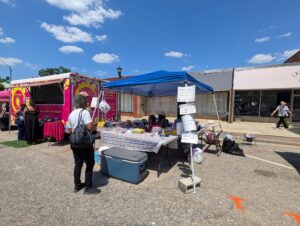 Outdoor street market with a food truck, people browsing under a blue tent, handwritten menu signs, and clear skies with scattered clouds above.