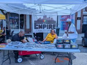 Three people at a street market booth selling t-shirts and merchandise under a white tent with an "EMPIRE" banner behind them.