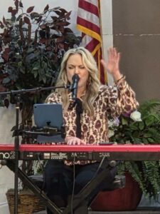 Woman singing and playing keyboard, raising one hand, in front of an American flag and plants, using a microphone and a tablet on a stand.