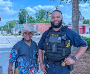 A smiling police officer stands next to a boy in a cowboy hat and Hawaiian shirt, with trees and police tape in the background on a sunny day.