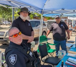 Three men, including a police officer in uniform, stand and sit under a blue tent at an outdoor event with tables and chairs.