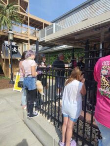 People, including children, stand by a black metal fence outside a building on a sunny day, some looking through the bars, with a pirate flag in the background.