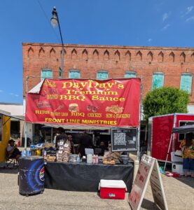 BBQ food stand with red banner offering ribs, turkey legs, chicken, and BBQ sauce, set up outdoors in front of a brick building under clear skies.