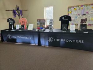 Merchandise table for The Browders with t-shirts, hats, and bags displayed in a church setting with cross and decorated bulletin board in the background.