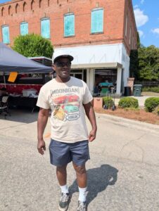 Man wearing a Hoonigan t-shirt, shorts, glasses, and cap, standing on a street near a red brick building and vendor tents on a sunny day.