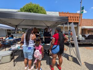People gather under a canopy tent at an outdoor event, talking at a table with pamphlets. Sunny day, brick buildings in the background.