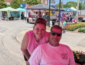 A smiling woman and a man in sunglasses pose together at an outdoor market with colorful tents and flowers in the background.