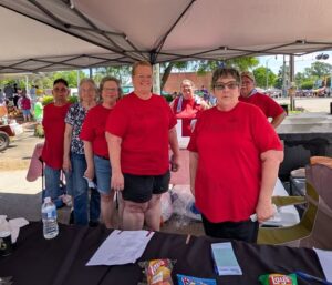 A group of people wearing red shirts stand under a canopy at an outdoor event, with snacks and papers on a table in front of them.
