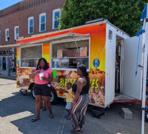 Three people, including a child, stand near a colorful food truck on a sunny day, with buildings and trees visible in the background.