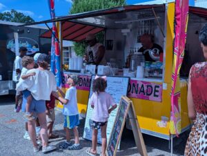 People, including children, stand in line at a bright yellow lemonade food truck on a sunny day, ordering drinks and interacting with the vendors inside.