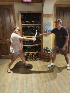 Two people playfully duel with toy swords indoors, standing on wooden floor near pirate hats, plush seals, and a "Mermaid Crossing" sign.