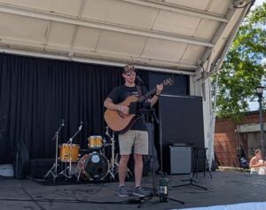 A man wearing sunglasses and a cap plays an acoustic guitar while singing on an outdoor stage with drums and microphones in the background.