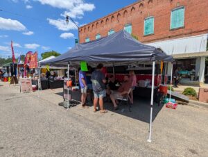 People gather under a tent at an outdoor market with food stalls and a brick building in the background on a sunny day.
