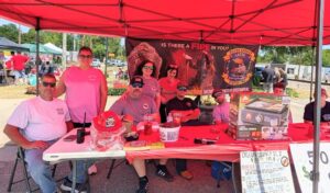 Group of people at a fire department booth under a red tent, with firefighting items, safety tips, and recruitment banner in the background at an outdoor event.