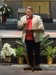 Man in beige blazer and red shirt speaks into a microphone at a podium, surrounded by potted plants and flowers on a stage.