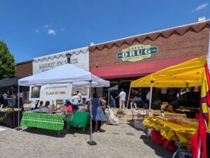 People gather at an outdoor market event under tents in front of Market on Main and Latta Drug Company on a sunny day.
