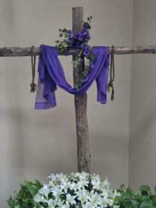 Wooden cross draped with purple cloth and flowers, surrounded by white lilies and green leaves, set against a plain background.