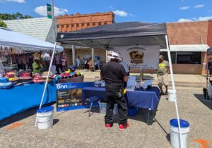 Outdoor street fair with vendor tents, hats for sale, and people browsing. A banner reads "Latimer Historic School Legacy." Bright sunny day with blue skies.