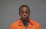 A man wearing an orange prison jumpsuit stands against a gray background, looking directly at the camera.