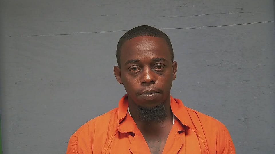 A man wearing an orange prison jumpsuit stands against a gray background, looking directly at the camera.