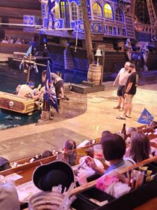 Audience watches a pirate-themed performance with actors near a mock ship and water, some wearing costumes, while others sit at tables with drinks and food.