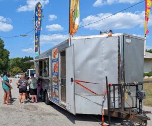 People stand in line at a silver food truck with colorful banners, selling hot dogs, corn dogs, and funnel cakes on a sunny day.